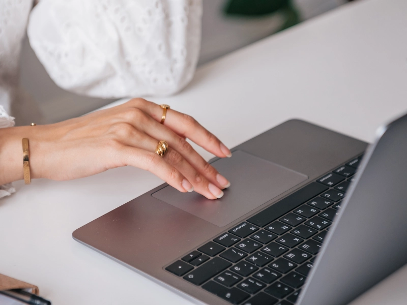 Das Bild zeigt eine Nahaufnahme einer Hand, die mit elegantem Goldschmuck verziert ist und auf dem Trackpad eines Laptops arbeitet. Die Person trägt ein weißes, leicht strukturiertes Oberteil, und der Laptop befindet sich auf einem weißen Schreibtisch.