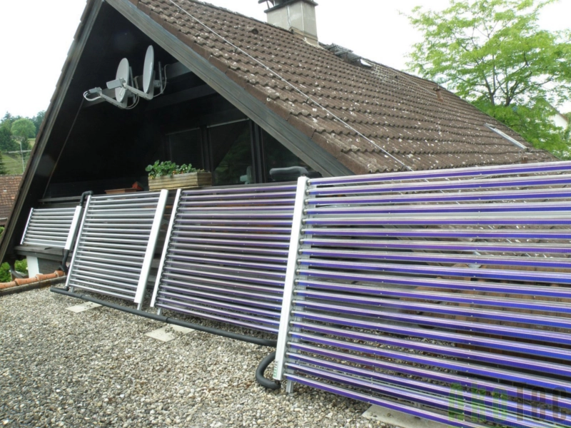 Solarthermieanlage mit Vakuumröhrenkollektoren auf einem Flachdach installiert, vor einem Haus mit Satteldach und Satellitenschüsseln. Effiziente Kombination von Solartechnologie mit bestehender Heizungsanlage zur nachhaltigen Energiegewinnung.