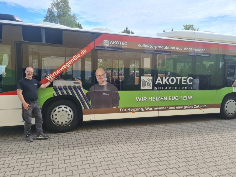 Werbebus von AKOTEC Solarthermie mit auffälligem Branding und Slogan 'WIR HEIZEN EUCH EIN!' für Heizung, Warmwasser und eine grüne Zukunft. Ein Mitarbeiter steht neben dem Bus, der für Kollektorproduktion aus Angermünde wirbt.