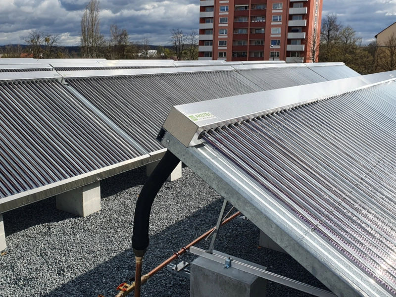 Großflächige Solarthermieanlage mit Vakuumröhrenkollektoren auf einem Flachdach, im Hintergrund ein Wohngebäude. Effiziente Nutzung von Sonnenenergie zur nachhaltigen Wärmeversorgung von Mehrfamilienhäusern.