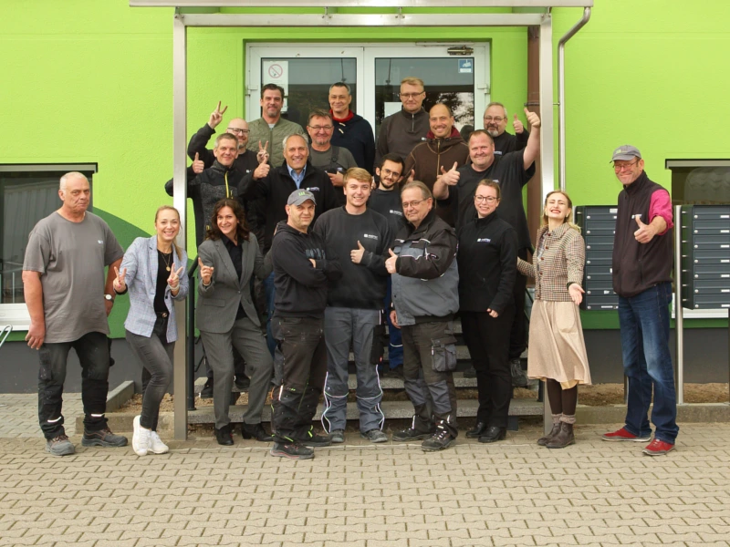 Teamfoto der AKOTEC-Mitarbeiter vor einem modernen Firmengebäude mit grüner Fassade. Die Gruppe zeigt Daumen-hoch-Gesten und symbolisiert ein motiviertes Team sowie einen zukunftsfähigen Arbeitgeber im Bereich Solarthermie.