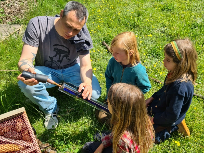 Erwachsener zeigt einer Gruppe von Kindern im Freien die Funktionsweise einer Solarthermie-Vakuumröhre. Projekttag zur Solarwärme an einer freien Draußenschule zur Förderung von Umweltbewusstsein und erneuerbaren Energien.