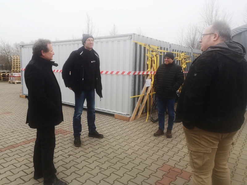 Gruppe von vier Männern im Gespräch vor einem Container mit gelben Befestigungselementen auf einem Firmengelände. Diskussion über MEGA-Kollektoren für Deutschlands größte Prozesswärme-Solarthermieanlage.