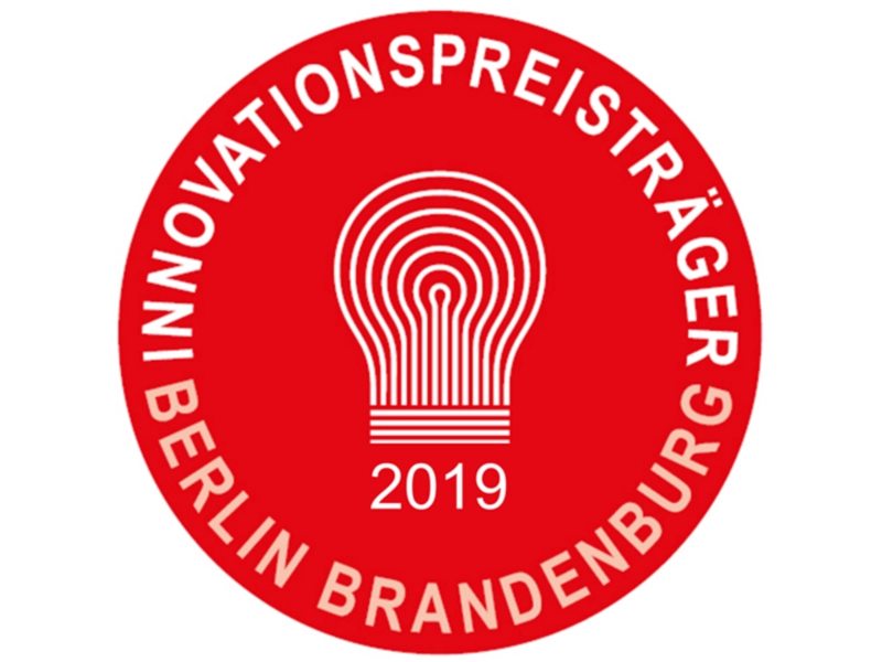 Das Bild zeigt das Logo des Innovationspreisträgers Berlin Brandenburg 2019 in Form eines roten Kreises. In der Mitte befindet sich ein stilisiertes Glühbirnen-Symbol, das für kreative Ideen und technologische Innovation steht, mit der Jahreszahl ‚2019‘ darunter. Der Schriftzug ‚INNOVATIONSPREISTRÄGER BERLIN BRANDENBURG‘ umrahmt das Logo.