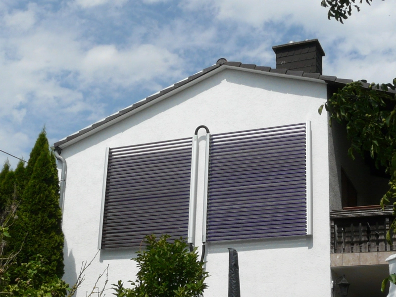 Das Bild zeigt die Fassade eines weißen Einfamilienhauses mit zwei großen, vertikal angebrachten Solarthermie-Kollektoren. Die Kollektoren sind symmetrisch auf der Hauswand montiert und durch ein zentrales Rohrsystem verbunden.