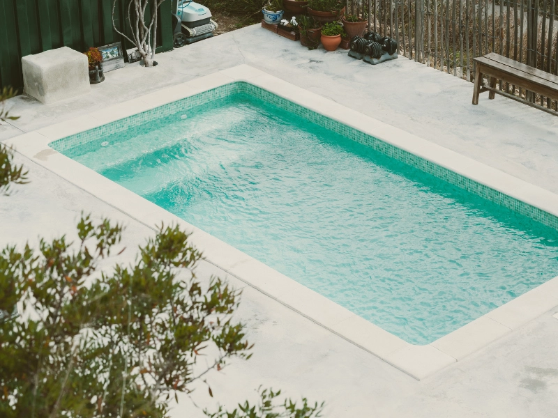 Das Bild zeigt einen rechteckigen, in den Boden eingelassenen Pool mit klarem, türkisfarbenem Wasser. Der Pool befindet sich in einem gepflegten Außenbereich mit einer Betonumrandung, umgeben von einigen Pflanzen, Töpfen und Gartenmöbeln.