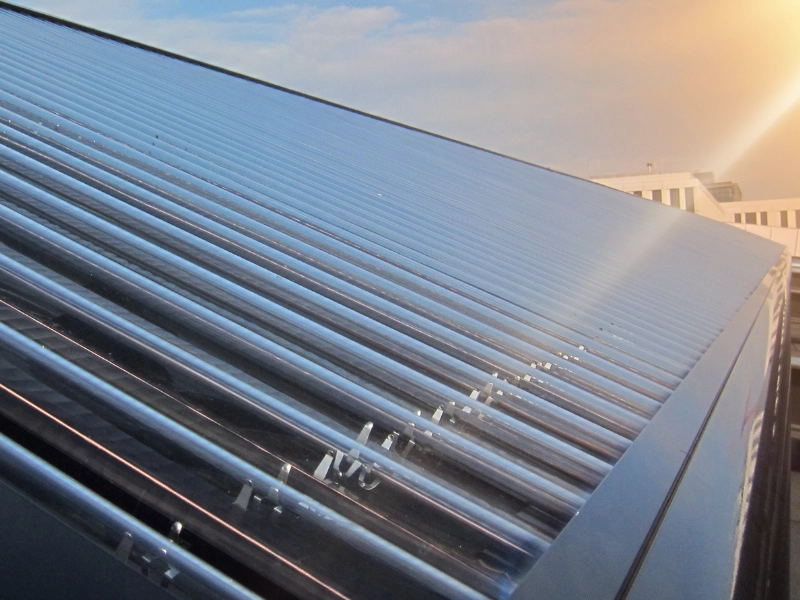 Nahaufnahme eines Solarthermie-Kollektors mit glänzenden Vakuumröhren, die schräg auf einem Dach montiert sind. Die Sonne reflektiert auf der glatten Oberfläche, wodurch ein Lichtreflex entsteht. Im Hintergrund sind unscharf moderne Gebäude und ein blauer Himmel mit leichten Wolken zu erkennen.