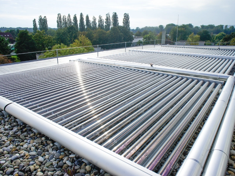 Das Bild zeigt eine großflächige Solarthermie-Anlage mit Vakuumröhrenkollektoren, die auf einem Flachdach mit Kiesbett installiert sind. Die Kollektoren sind in Reihen angeordnet und von einem stabilen Montagesystem eingerahmt.