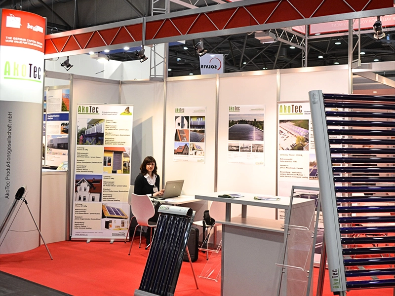 Messestand von AKOTEC SOLARTHERMIE mit Informationsmaterial, Solarthermie-Kollektoren und einer Mitarbeiterin am Laptop.