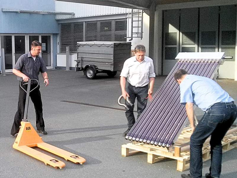 Drei Männer bewegen einen Vakuumröhrenkollektor auf einer Holzpalette mit einem Hubwagen vor einem Industriegebäude. Szene zeigt den Transport von Solarkollektoren für AKOTEC SOLARTHERMIE.