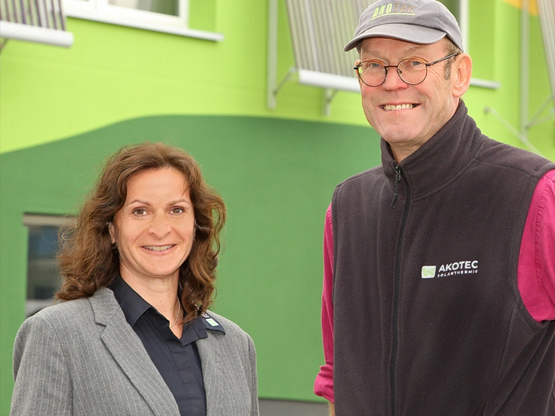 Zwei Personen vor einem grünen Firmengebäude der AKOTEC SOLARTHERMIE. Die Frau trägt einen grauen Blazer, der Mann eine AKOTEC-Weste und eine Kappe mit Logo, beide lächeln in die Kamera.