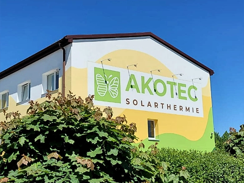 Akotec Solarthermie Firmengebäude mit grün-gelber Fassade und großem Logo, umgeben von grünen Büschen und strahlend blauem Himmel – 15 Jahre Akotec Jubiläum.