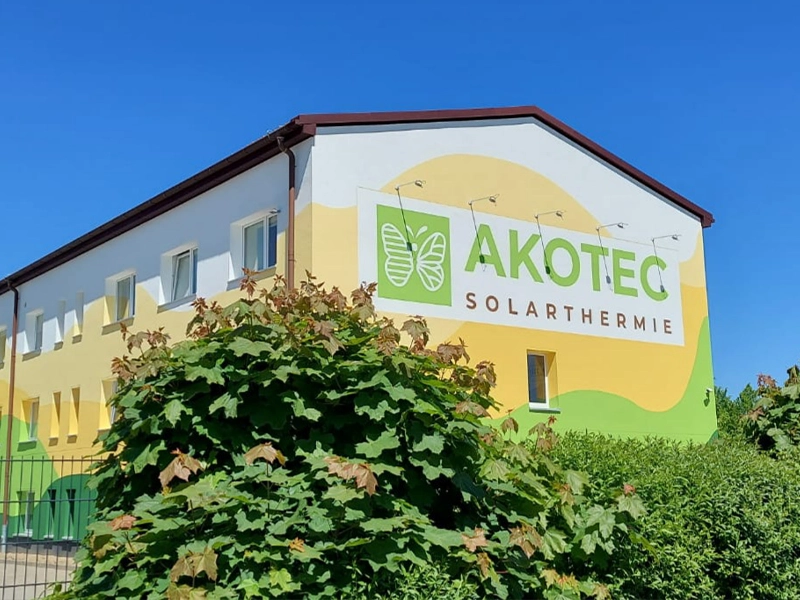 Gebäude von AKOTEC SOLARTHERMIE mit grün-gelber Fassade, großem Firmenlogo und Schmetterlingssymbol an der Wand, umgeben von Büschen und blauem Himmel.