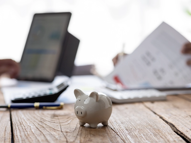 Weißes Sparschwein im Vordergrund, im Hintergrund unscharf Personen mit Finanzdokumenten und Laptop. Symbolisiert alternative Finanzierungsmöglichkeiten für Solarthermie-Anlagen.