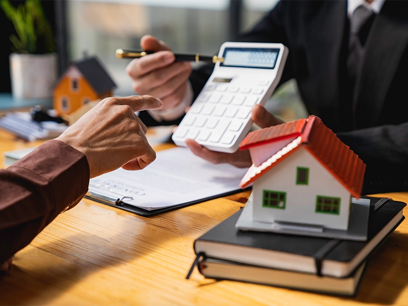 Zwei Personen diskutieren über Finanzierungen, eine hält einen Taschenrechner, die andere zeigt mit dem Finger darauf. Im Vordergrund ein Modellhaus mit rotem Dach, symbolisierend für die Förderung von Solarthermie-Anlagen.