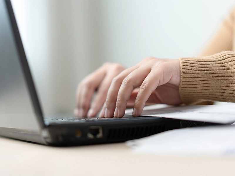 Nahaufnahme von Händen, die auf der Tastatur eines Laptops tippen. Im Vordergrund liegen unscharf einige Blätter auf dem Tisch. Die Person trägt einen beigefarbenen Pullover.