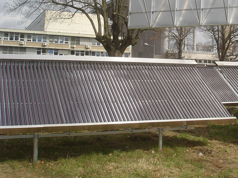 Große Solarthermie-Kollektoren sind auf einer Freifläche installiert. Die Kollektoren bestehen aus zahlreichen Vakuumröhren, die in einem Metallrahmen fixiert sind. Im Hintergrund sind ein Gebäude mit vielen Fenstern sowie Bäume ohne Blätter sichtbar. Der Himmel ist teils bewölkt.