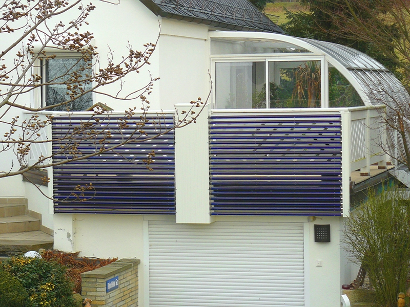 Moderne Solarthermie-Installation an einem Balkon eines weißen Einfamilienhauses. Die blauen Vakuumröhren sind horizontal angebracht und dienen sowohl der Energiegewinnung als auch als Sichtschutz. Im Hintergrund ist ein Wintergarten mit Glasdach zu sehen, daneben ein Baum mit kahlen Ästen.