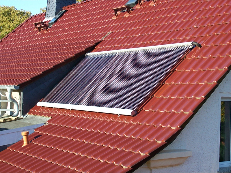 Rotes Ziegeldach eines Einfamilienhauses mit einem installierten Solarthermie-Kollektor. Der Kollektor besteht aus mehreren parallelen Vakuumröhren, die in einem Winkel zur optimalen Sonneneinstrahlung angebracht sind. Im Hintergrund sind weitere Dachflächen mit Dachfenstern und ein Teil eines Schornsteins zu sehen.