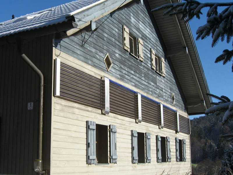 Traditionelles, zweistöckiges Holzhaus mit blauen Fensterläden und geschlossenen Klappläden im oberen Bereich. An der Fassade sind mehrere vertikale Solarthermie-Kollektoren montiert. Das Dach ist teilweise mit Schnee bedeckt, und im Hintergrund sind schneebedeckte Bäume sichtbar, was auf eine winterliche Umgebung hinweist.