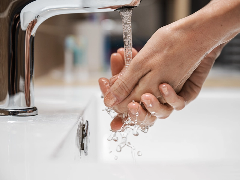 Nahaufnahme von Händen, die unter fließendem Wasser aus einem modernen Wasserhahn gewaschen werden. Wassertropfen spritzen in das weiße Waschbecken, während der Hintergrund leicht unscharf ist. Symbolisch für die Nutzung von Warmwasser im Haushalt.