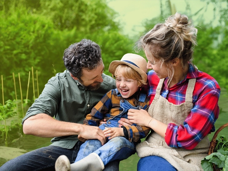 Glückliche Familie im Garten: Ein Mann, eine Frau und ein lachendes Kind sitzen zusammen im Grünen. Der Mann trägt ein grünes Hemd, die Frau ein kariertes Hemd mit einer Schürze, und das Kind hat einen Strohhut und Latzhosen an. Die Familie lacht und genießt die gemeinsame Zeit inmitten von üppigem, grünem Hintergrund.