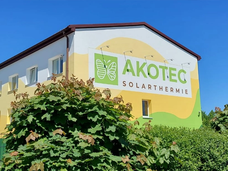 Gebäude der Firma AKOTEC SOLARTHERMIE mit farbenfroher Fassade in Gelb- und Grüntönen, großem Logo und Schriftzug an der Wand. Im Vordergrund grüne Sträucher, blauer Himmel im Hintergrund.