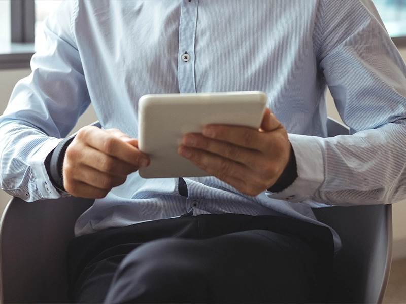 Ein Mann in einem hellblauen Hemd sitzt auf einem Stuhl und hält ein Tablet mit beiden Händen. Das Tablet wird im Querformat gehalten. Der Fokus liegt auf dem Tablet und den Händen, das Gesicht des Mannes ist nicht sichtbar. Im Hintergrund ist ein heller, unscharfer Innenraum erkennbar.