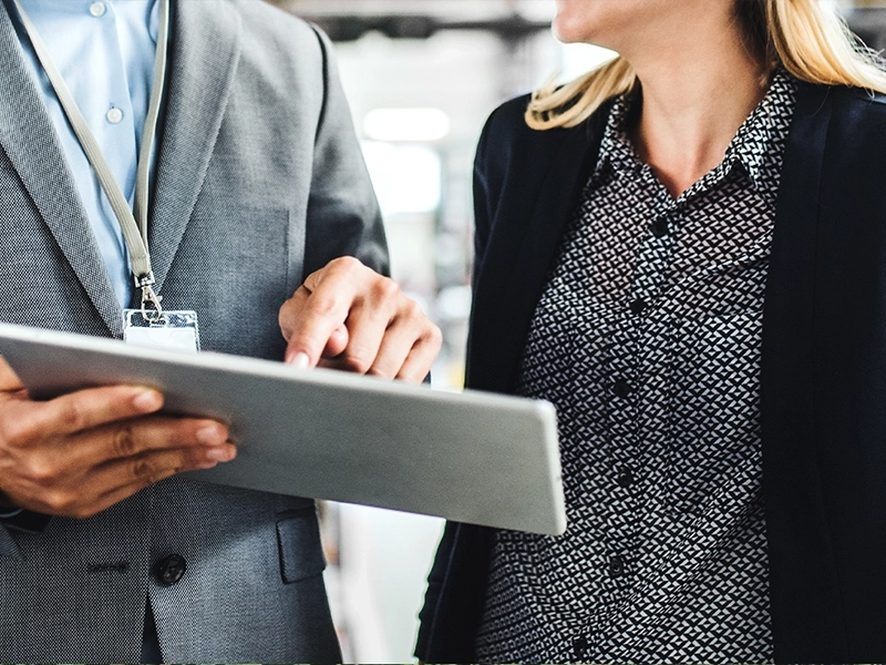 Zwei Geschäftspersonen in Business-Kleidung besprechen Informationen auf einem Tablet. Eine Person zeigt mit dem Finger auf den Bildschirm, während die andere aufmerksam zuhört.