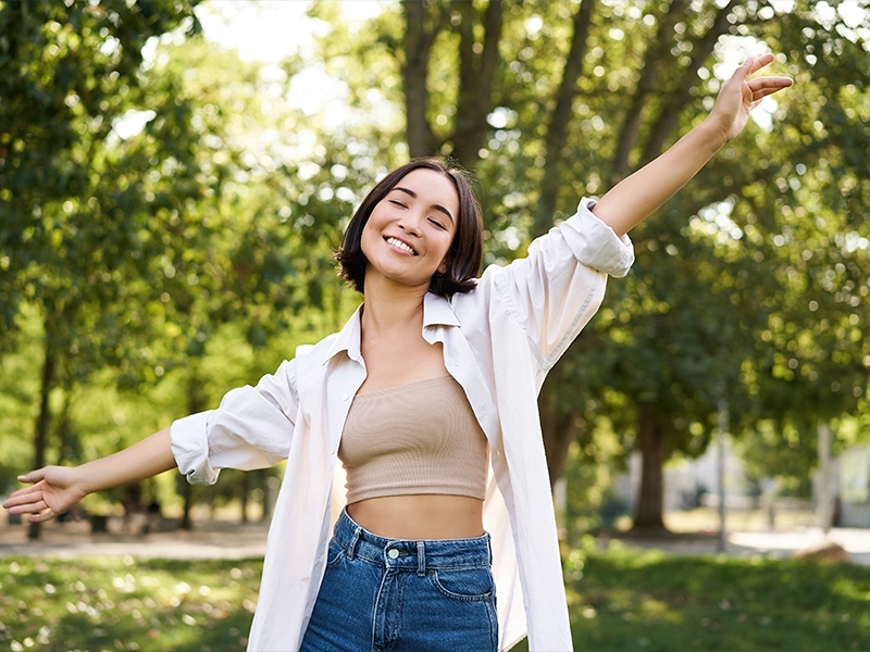 Junge, lachende Frau mit ausgebreiteten Armen im Park. Sie trägt ein beiges Top, eine offene weiße Bluse und Jeans, im Hintergrund grüne Bäume und Sonnenlicht.