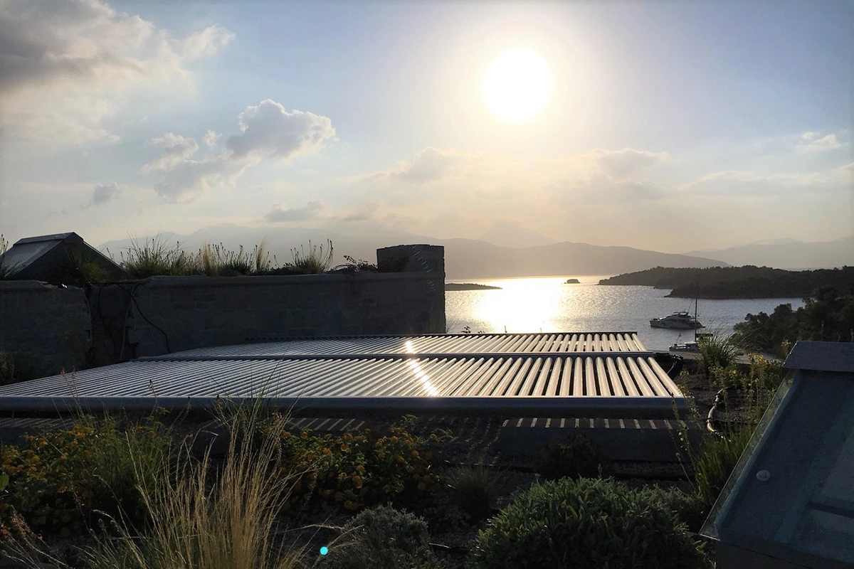 Solaranlage auf einem begrünten Dach im Sonnenuntergang, im Hintergrund ein ruhiges Meer mit Booten und hügeliger Landschaft.