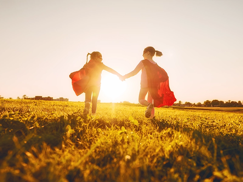 Zwei Kinder mit roten Umhängen laufen Hand in Hand über eine sonnige Wiese, Symbol für eine nachhaltige und strahlende Zukunft.