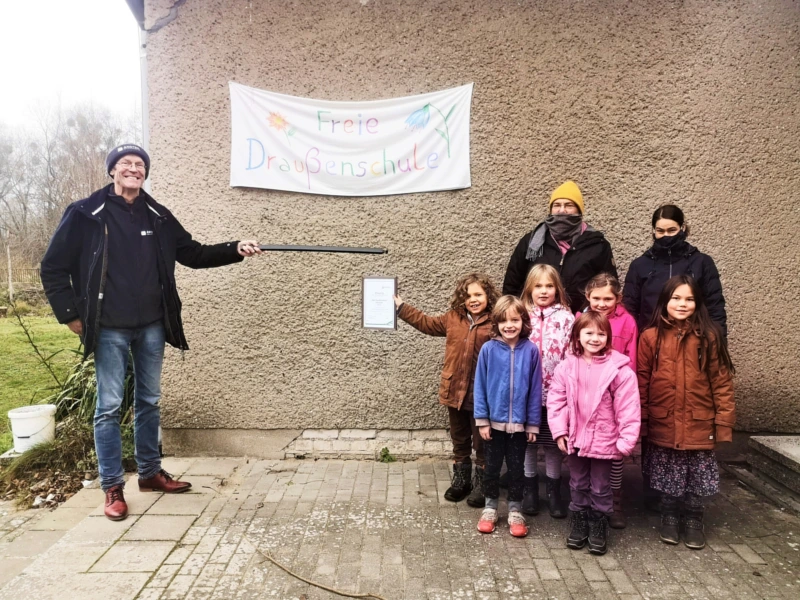 Gruppe von Kindern und Erwachsenen vor einem Gebäude mit einem Banner der 'Freien Draußenschule'. Ein Mann hält eine Urkunde, symbolisch für die Unterstützung der Schule durch AKOTEC im Bereich nachhaltiger Bildung und Umweltprojekte.