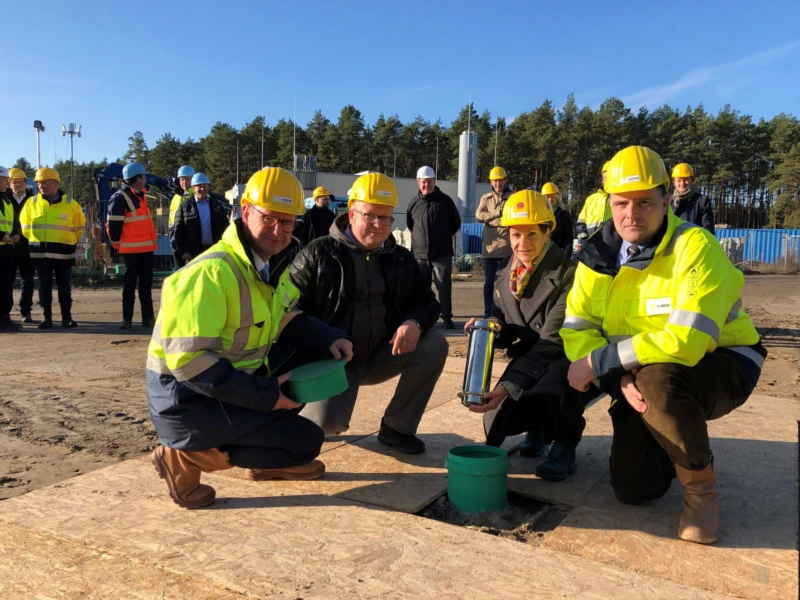 Grundsteinlegung mit vier Personen in Schutzkleidung und gelben Helmen, die eine Zeitkapsel in eine vorbereitete Öffnung einsetzen. Im Hintergrund weitere Bauarbeiter auf einer Baustelle, symbolisch für den Start eines Infrastrukturprojekts zur Versorgung der Erdgasstraße Eugen in Kienbaum.