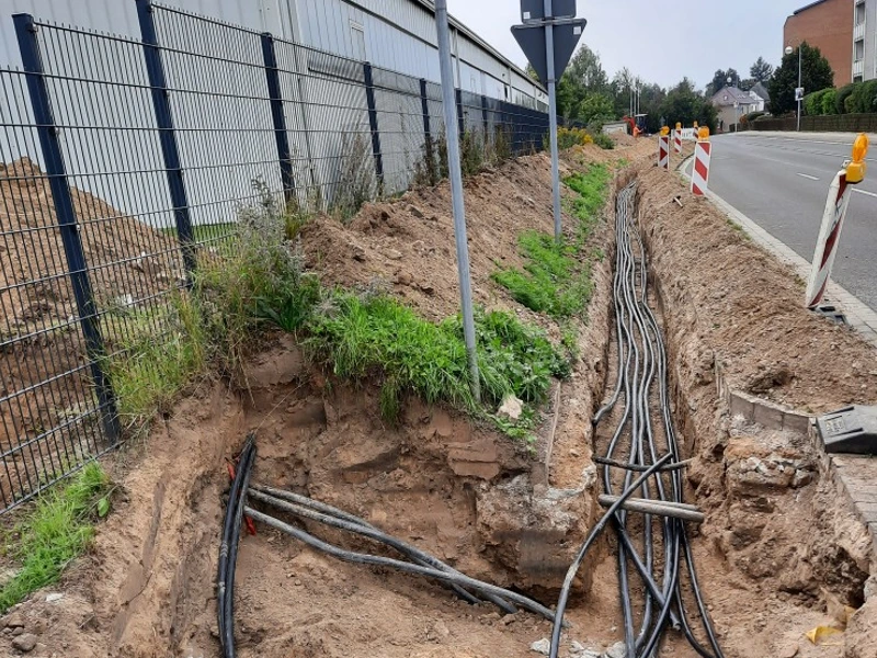 Baustelle mit geöffnetem Kabelgraben entlang einer Straße, zahlreiche verlegte Versorgungsleitungen sichtbar. Symbol für Infrastrukturentwicklung im Rahmen von Hallenerweiterungsprojekten für zukünftiges Wachstum.