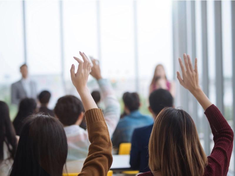 Teilnehmer einer Weiterbildung oder Schulung in einem modernen Seminarraum mit großen Fenstern, mehrere Personen heben die Hand, um Fragen zu stellen. Fokus auf aktive Teilnahme und Wissensaustausch.